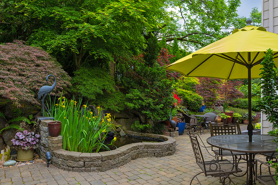 Home backyard hardscape and lush plants landscaping with garden furniture on paver brick patio.