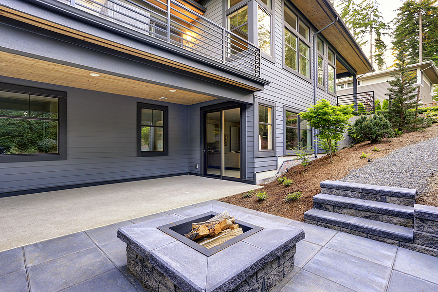 New modern home features a backyard with covered patio accented with a wood plank ceiling and a rectangular fire pit made of concrete and slate tiles. 