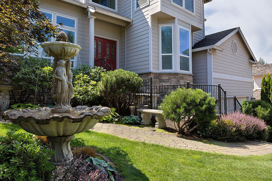 Residential single home in North America suburbs frontyard with manicured garden green lawn paver brick walkway bench and stone water fountain.