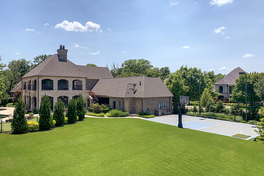 Outdoor shot of luxury house in suburban neighborhood with trees, bushes, lawn and basketball court on the backyard, blue sky background. 